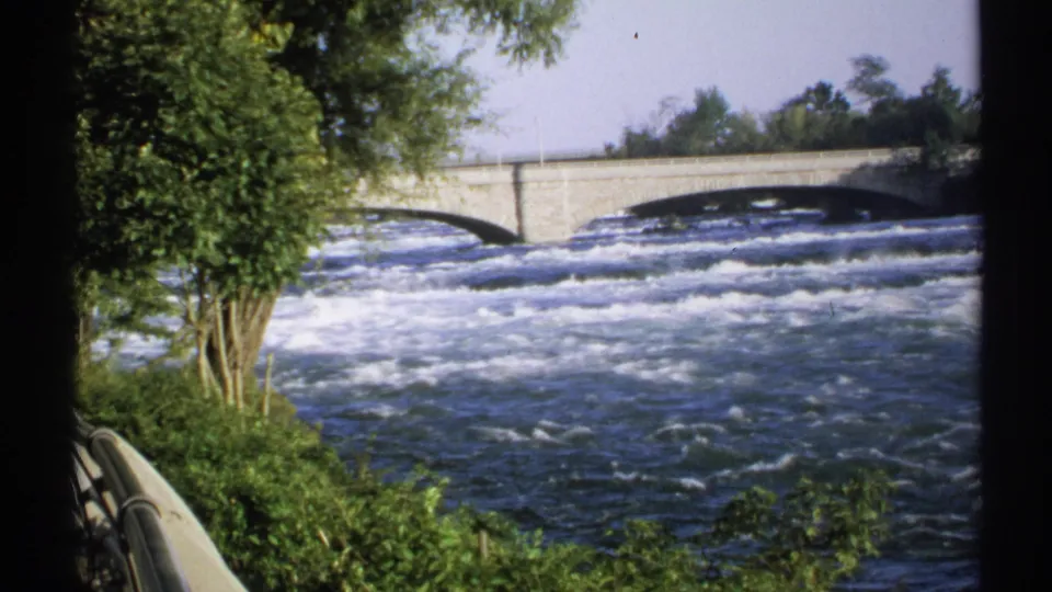 Fast water moves below a stone bridge in a New York 1971 home-movie view, with trees at the riverbank and film-edge shadows visible.
