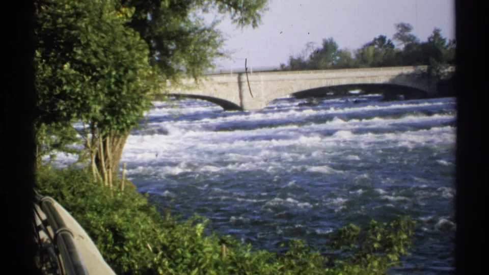 A 1971 New York 8mm frame shows broad whitewater, a stone bridge, green riverside trees, and a curved railing from a visitor overlook.