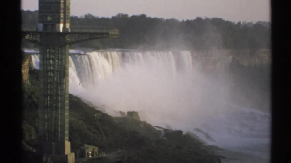 1971 8mm footage frame showing Niagara Falls, rising mist, a foreground tower structure, and a wooded rim in New York.