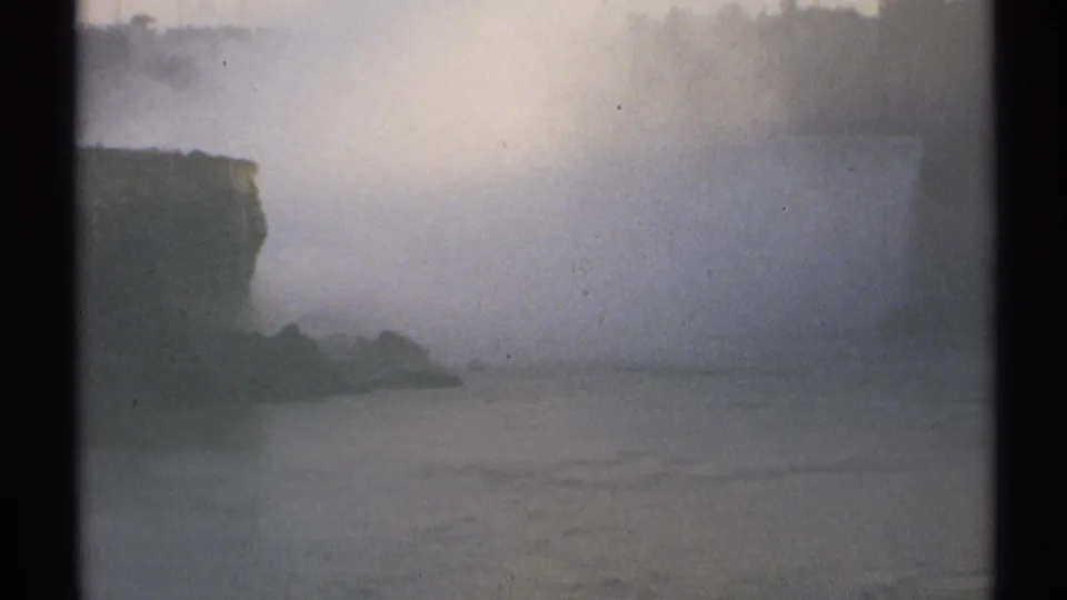 Misty 1971 Niagara Falls frame showing a rocky edge, obscured falling water, river surface, treeline, and dark gate borders.