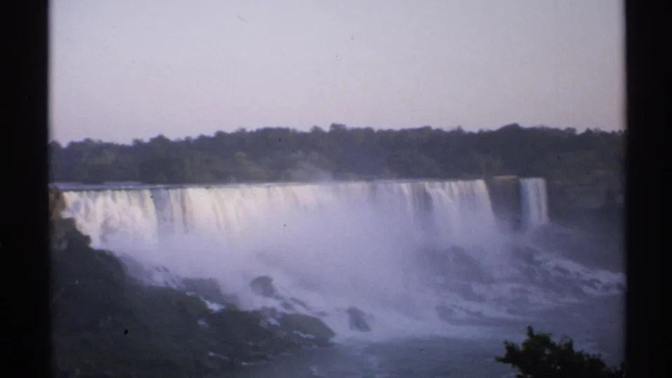 1971 8mm wide view of Niagara Falls with a bright cascade, rising mist, rocky base, green treeline, and visible film gate edges.