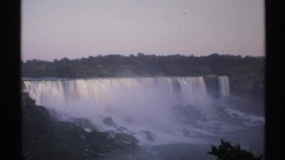 1971 Niagara Falls home-movie frame showing the broad cascade, heavy spray, rocky foreground, treeline, pale sky, and 8mm borders.