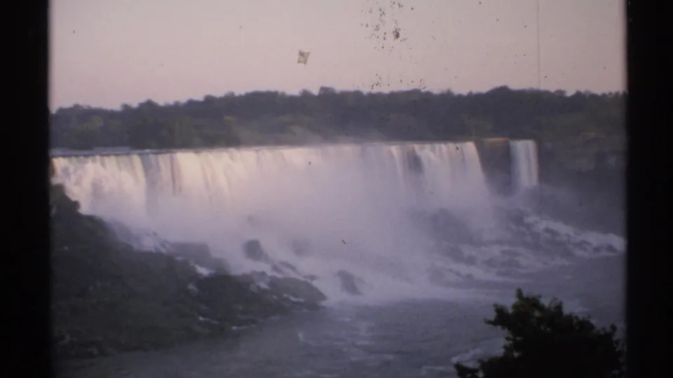 1971 8mm frame of Niagara Falls with bright cascade, misty lower gorge, trees on the horizon, and visible dust on the film.