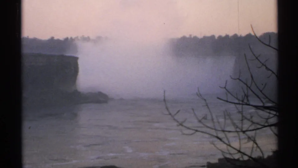 1971 Niagara Falls view with a large mist plume, cliff edge, river below, distant treeline, and foreground bare branches.