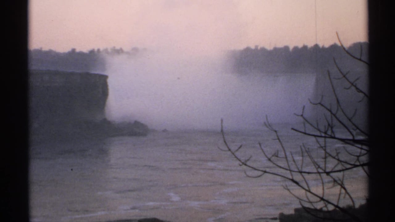 Distant waterfall mist behind a cliff, river water, and bare branches in foreground.