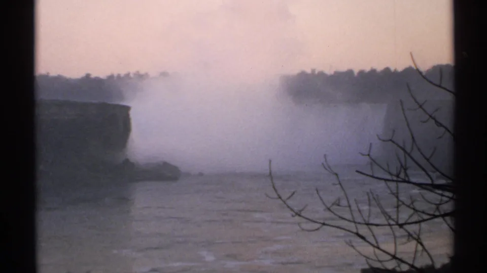1971 8mm frame of Niagara Falls with dense mist, dark rocky edge, foreground branches, and muted dusk-like color.