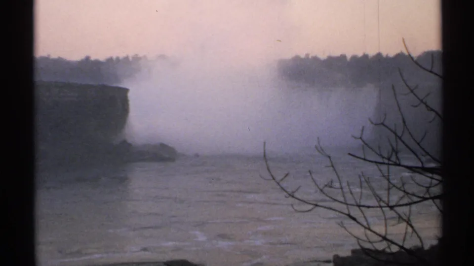 1971 New York home-movie view of Niagara Falls, with mist drifting over the river and dark branches in the foreground.