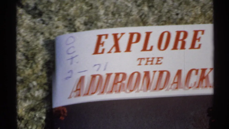Cropped 1971 New York home-movie frame showing the Explore the Adirondacks brochure title and handwritten marks on stone.