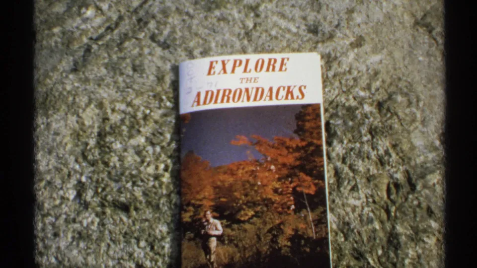 Wider 8mm frame of the Explore the Adirondacks brochure on stone, showing autumn foliage cover art and a small hiker figure.