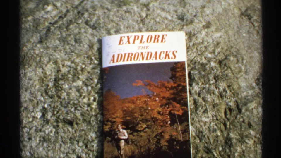 Wider 1971 8mm frame showing the Explore the Adirondacks brochure resting on textured stone with autumn landscape artwork.