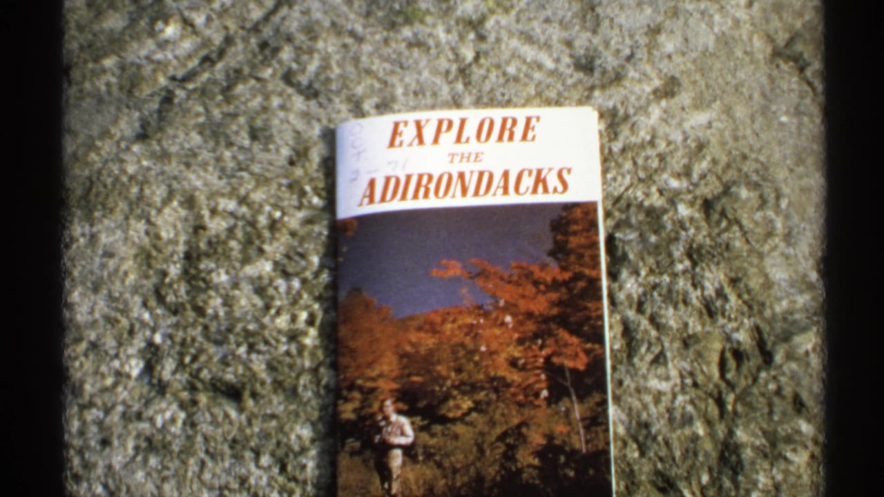 A 1971 Adirondacks travel brochure appears on a rough stone surface, with autumn foliage and a hiker printed on its cover.