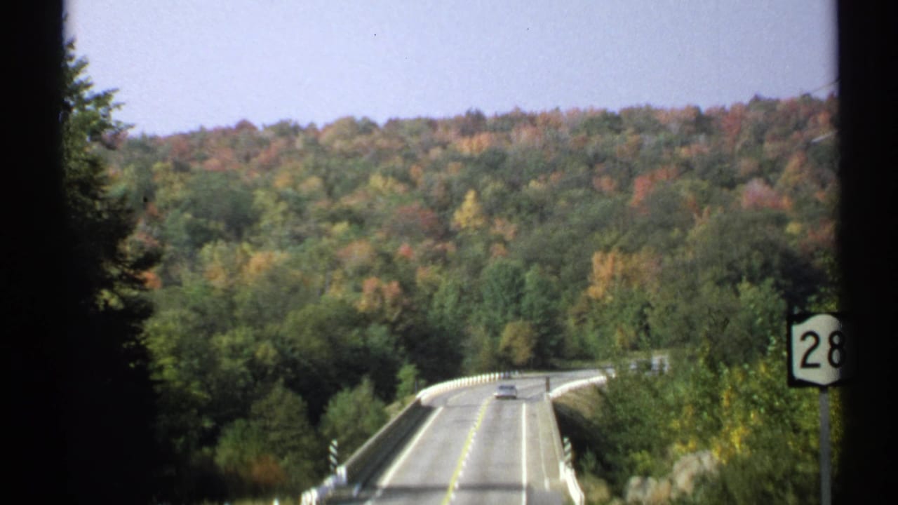 Two-lane road crossing a small bridge toward wooded hills in autumn color.