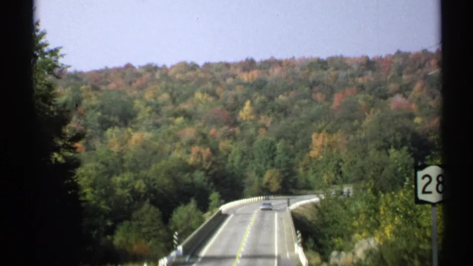 A two-lane Adirondack-area road runs over a small bridge toward wooded hills with early autumn color and a Route 28 sign at right.