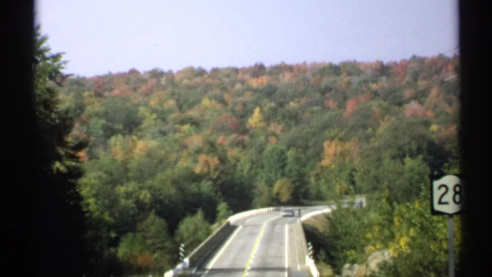The moving-vehicle view shows a Route 28 sign near a bridge, with layered green and orange wooded hills in New York.