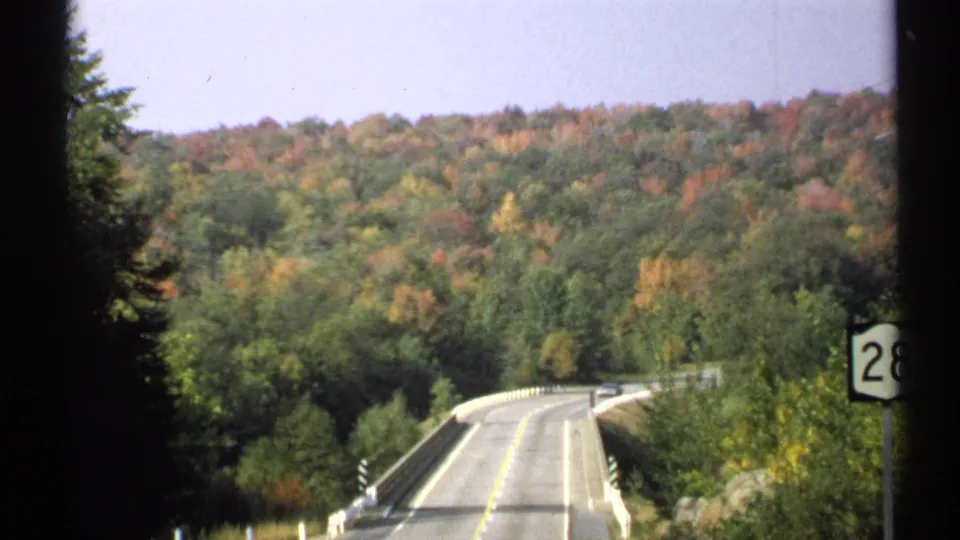 A scenic road over a bridge curves into Adirondack-area woods, with a Route 28 sign partly visible along the right edge.