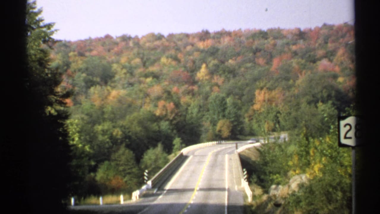 1971 New York 8mm frame showing a curving rural road, bridge rail, autumn woods, and dark vehicle-window edges.