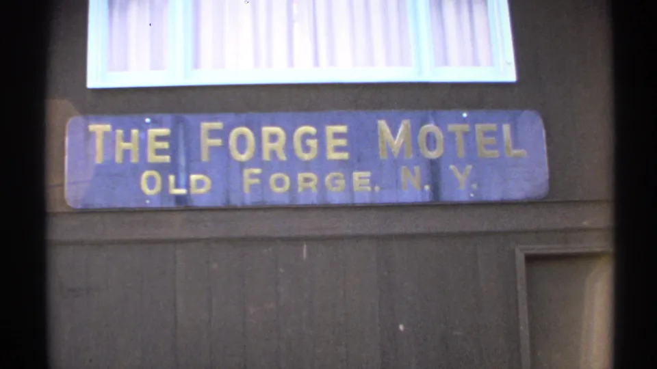 1971 Old Forge, New York close-up of The Forge Motel sign, with gold lettering, dark siding, and a partial doorway at the edge.