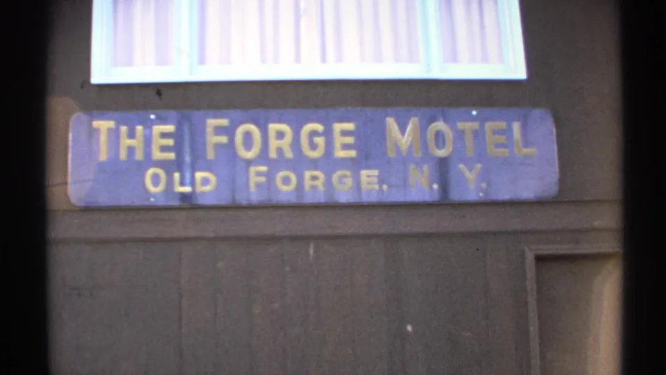 1971 Old Forge, New York frame of the blue Forge Motel sign below a bright window with vertical blinds on a dark wooden facade.