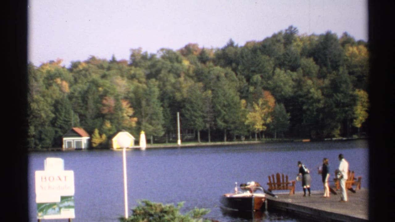 1971 New York home-movie frame showing people preparing near a docked motorboat on a wooded lake with a BOAT Schedule sign visible.
