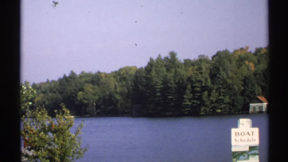 1971 New York home-movie view across a calm lake toward dense trees, with a partial BOAT Schedule sign and shoreline structure visible.