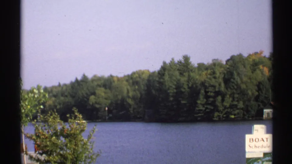 1971 New York lakeside view with evergreen trees, a boat schedule sign near shore, and small docks across the water.