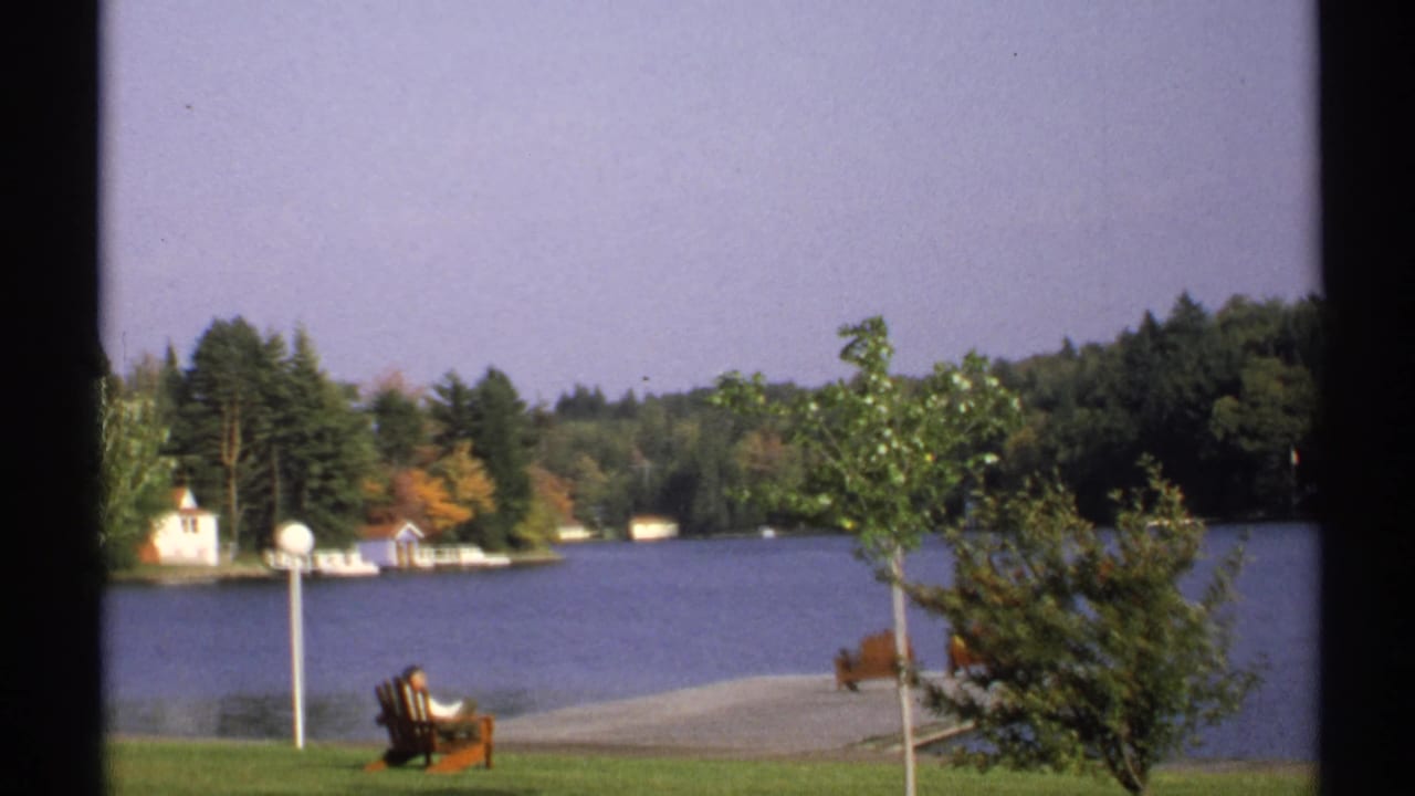 Person seated in an Adirondack chair beside a lake with trees and small buildings across the water.