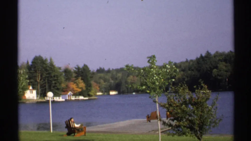 1971 New York home-movie frame showing chairs on a grassy lakeshore, a small tree, docks, and wooded shoreline buildings.
