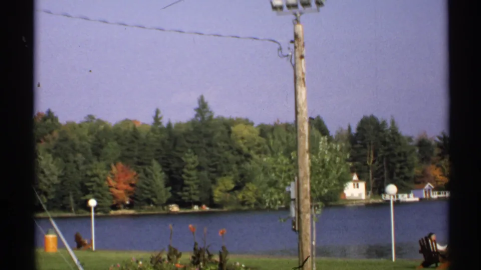 A 1971 New York lakeshore view includes a utility pole, lamps, Adirondack seating, wooded shoreline, and small waterfront buildings.