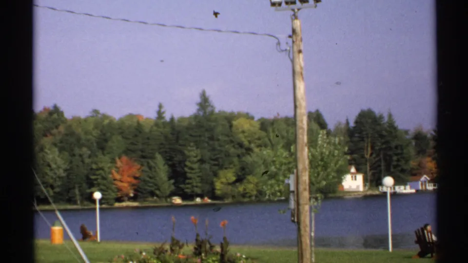 1971 New York 8mm frame showing a lakeside view with autumn trees, utility wires, lawn lamps, and a small white building across the water.
