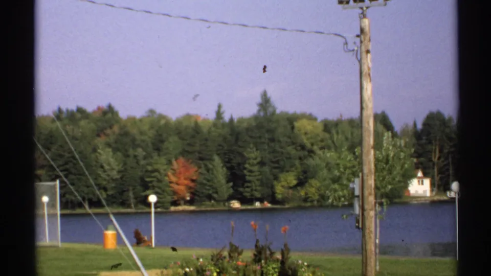 1971 New York home-movie frame of a quiet lakefront lawn, utility pole, overhead wires, and wooded shoreline with a small building beyond.