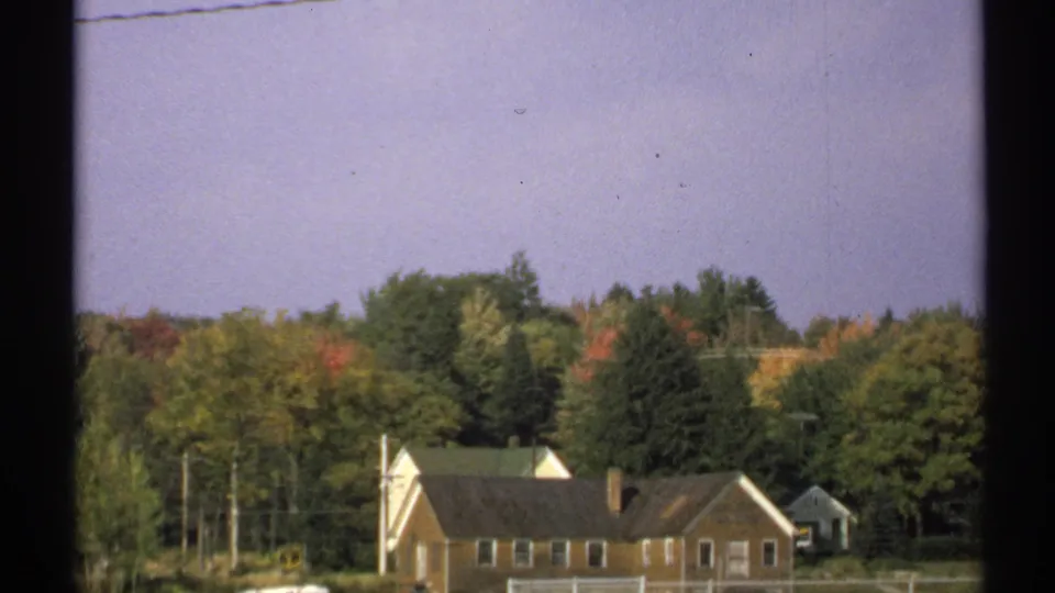 1971 New York 8mm frame showing low residential buildings near a wooded area, with autumn foliage and utility poles in view.