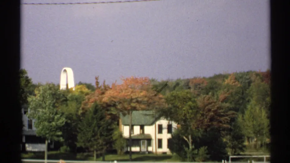 1971 New York 8mm frame with a white house, green roof, autumn-colored trees, pale sky, and dark film or window edge at left.