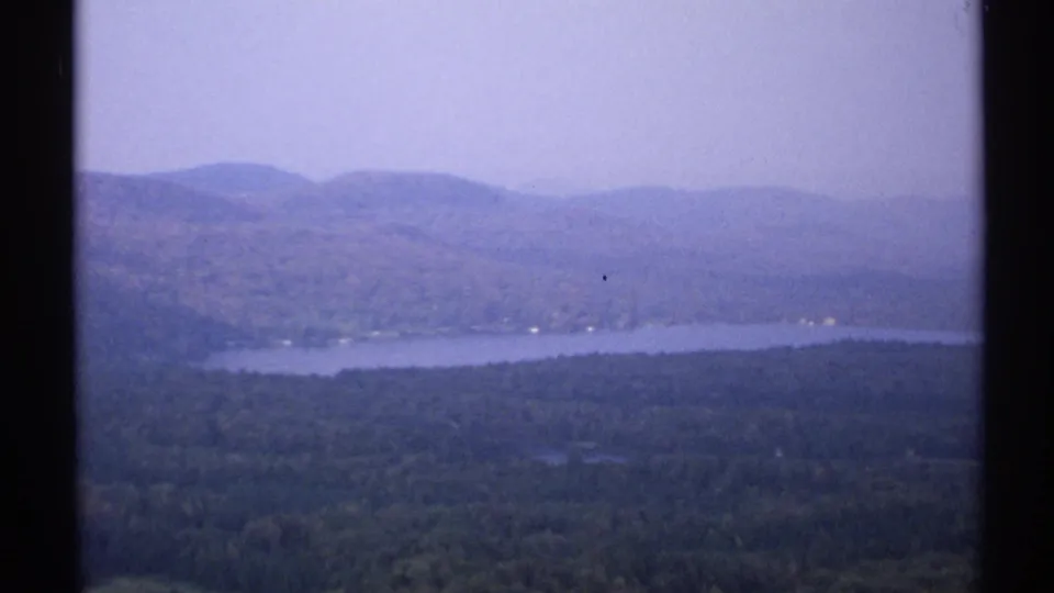 1971 New York 8mm landscape showing a calm lake, forested slopes, and distant ridges under a pale sky.