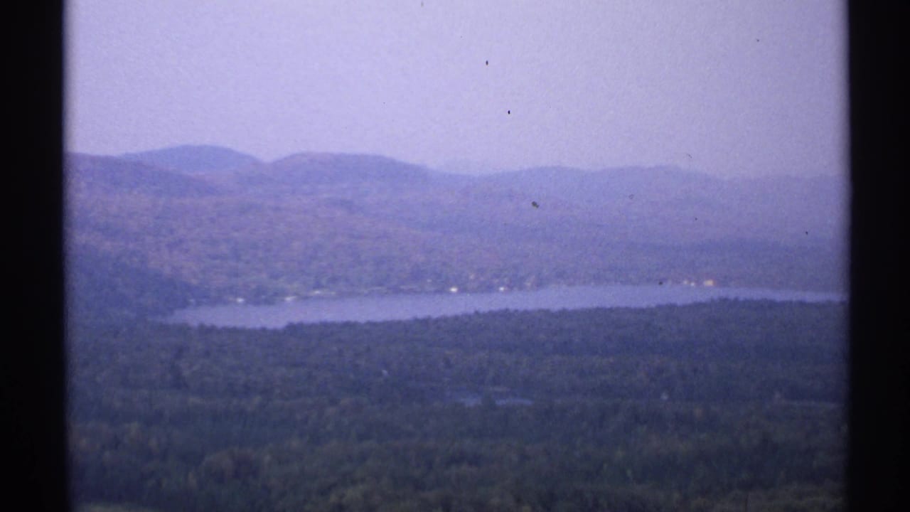 1971 New York 8mm view of a broad lake, wooded hills, and hazy distant mountains framed by dark film edges.