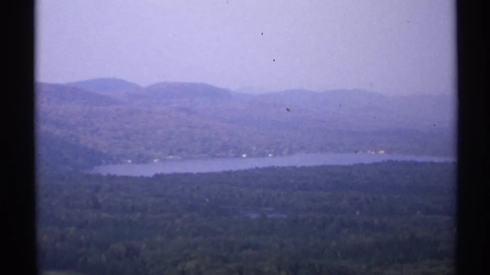 1971 New York 8mm view of a broad lake, wooded hills, and hazy distant mountains framed by dark film edges.