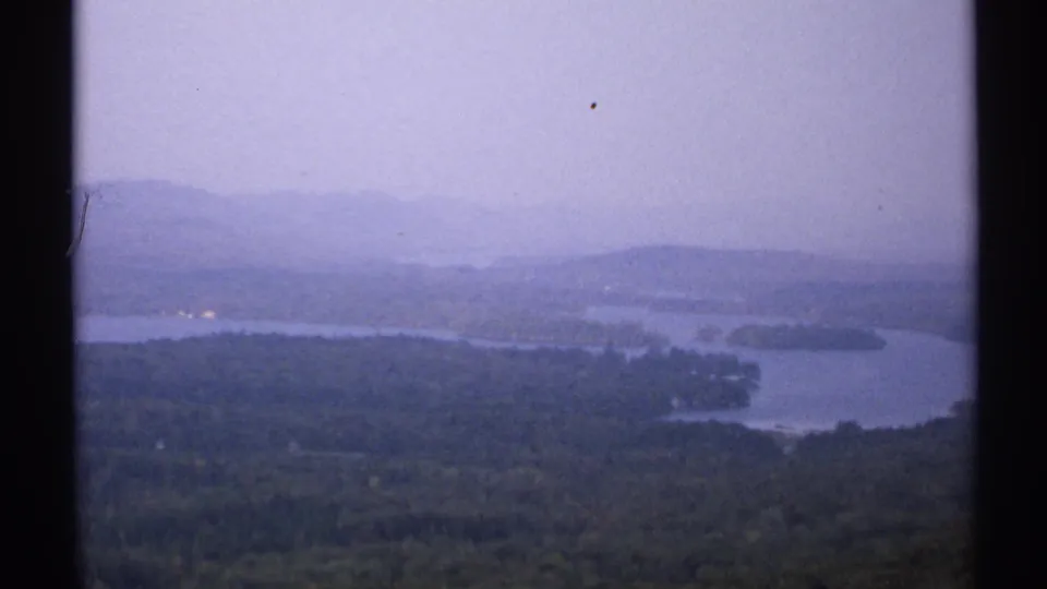 1971 New York 8mm frame showing a lake with wooded shorelines, small landforms, distant hills, and soft haze.