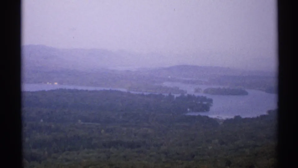 1971 New York home-movie frame of a hazy lake panorama with forest below, distant hills, and muted blue-purple film color.
