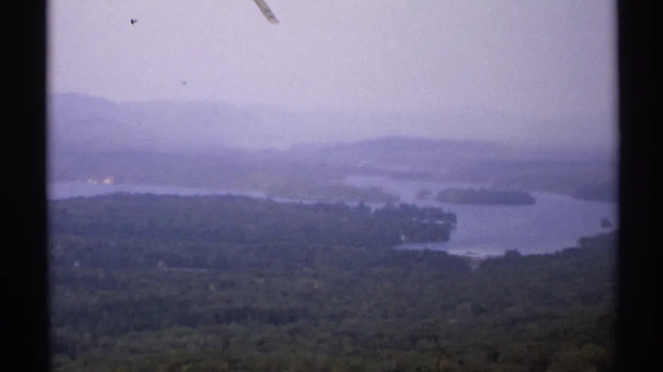 1971 New York 8mm view of a lake, wooded peninsulas, distant mountains, and visible film dust in a soft hazy frame.