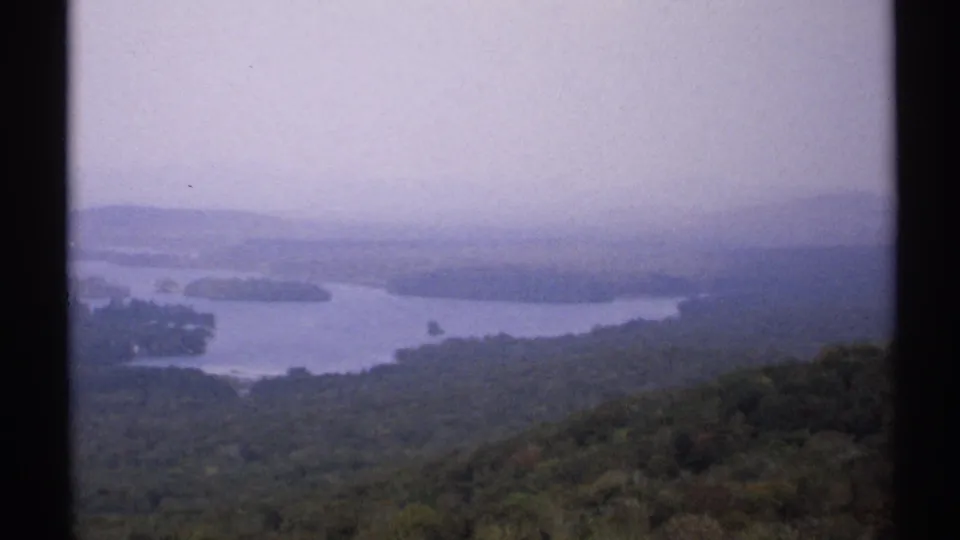 1971 New York 8mm frame showing a hazy lake panorama from a wooded mountain overlook, bordered by the film gate.