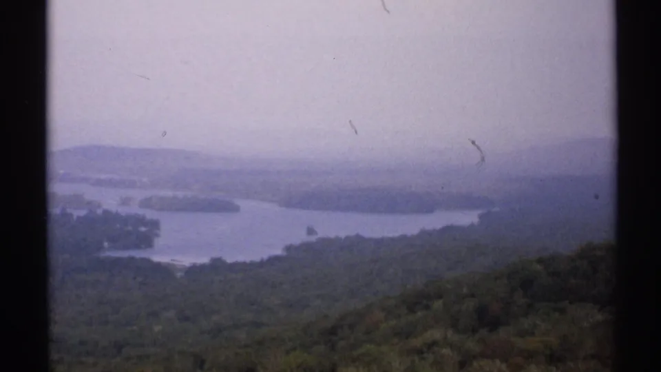 1971 New York home-movie frame of a wide lake and forested slopes viewed from an elevated mountain position.