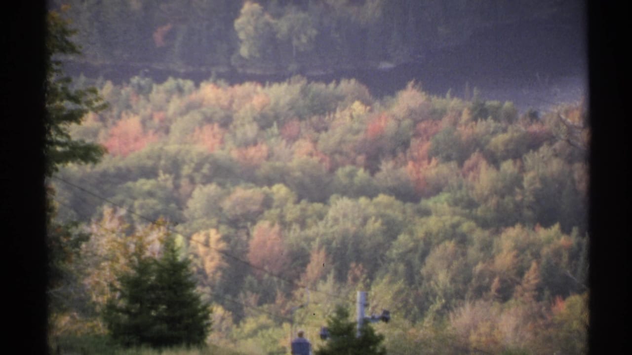 1971 New York 8mm frame showing a forested autumn hillside with a small distant person and cable car or lift equipment low in the view.