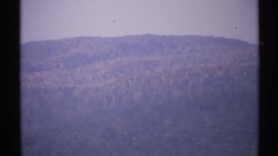 1971 New York home-movie frame of a distant autumn ridge under pale sky, with soft grain and dark gate edges.
