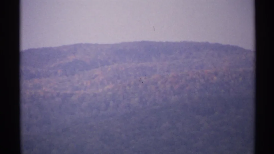 1971 New York home-movie frame of a wooded ridge in hazy light, with subdued purple and orange color shifts.