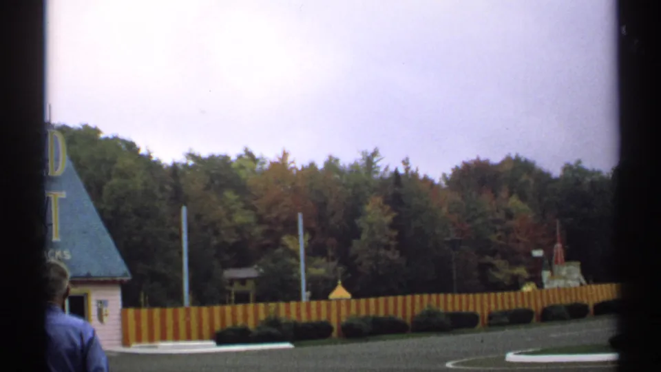1971 New York 8mm frame showing a man in a blue shirt near the Enchanted Forest grounds, with fence, trees and balloon-like object behind.