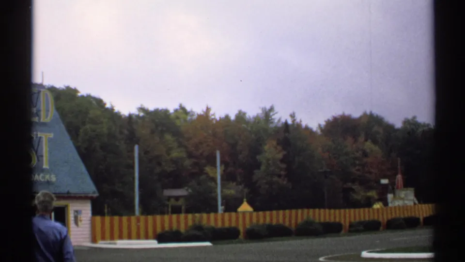 1971 New York frame with an adult visitor in the foreground and the Enchanted Forest grounds, striped fencing and trees behind him.