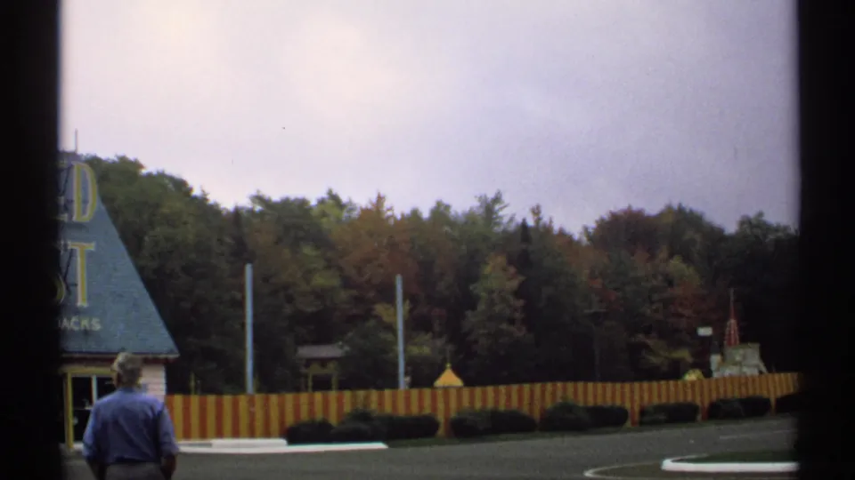 1971 New York frame with a man viewed from behind beside a blue-roofed stand, partial yellow lettering, red and yellow fence, hedges, and trees.