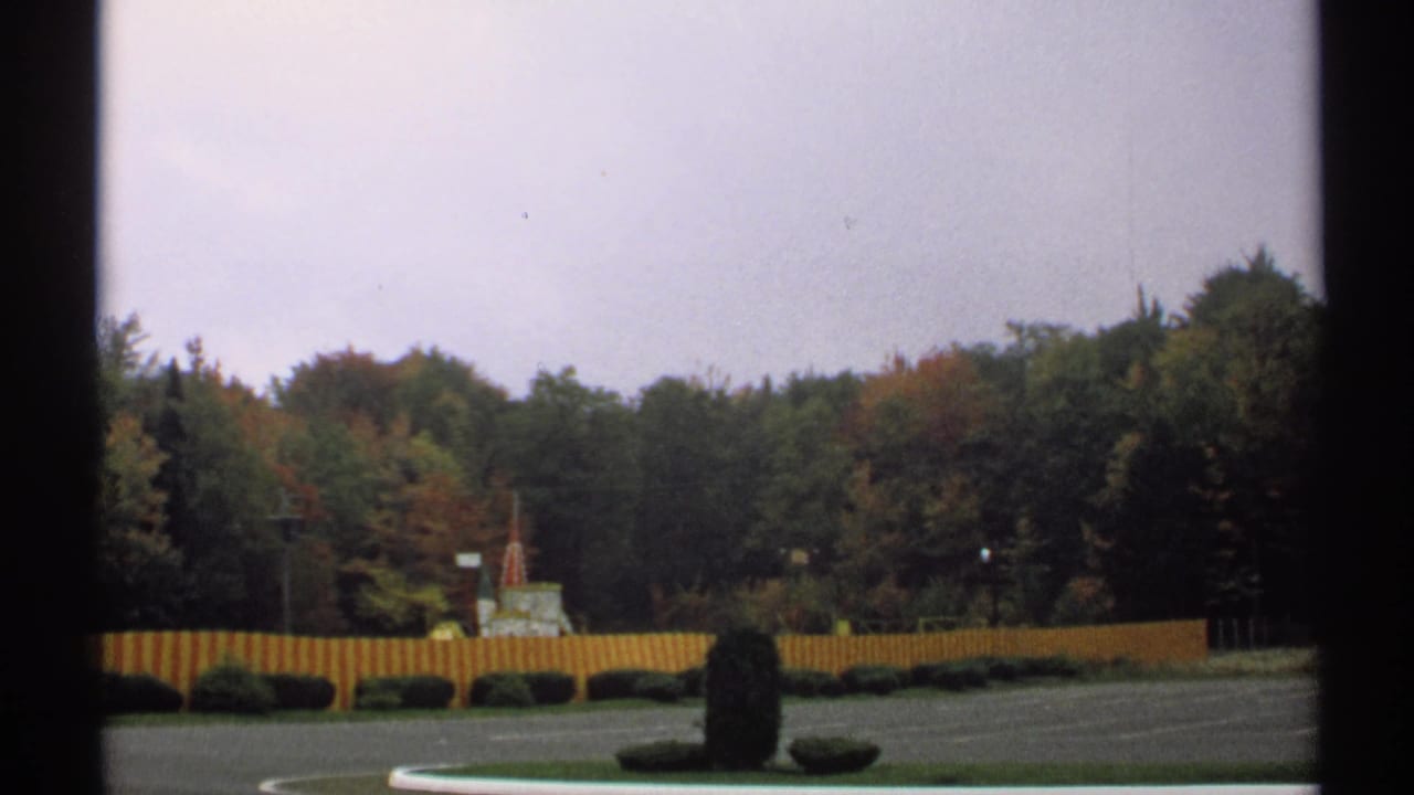 1971 New York home-movie view of a landscaped paved area with a red and yellow fence, clipped hedges, a small distant structure, and autumn-tinted trees.
