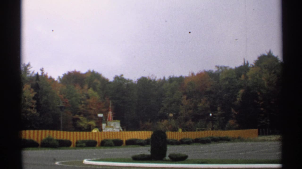 1971 New York frame showing a quiet landscaped outdoor area with red and yellow fencing, dark hedges, paved ground, and dense early autumn trees.