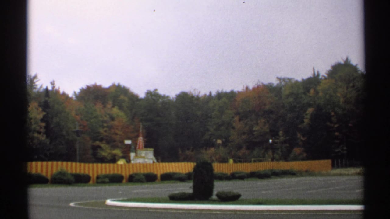 Roadside landscaped area with fence, shrubs, trees, and overcast sky.