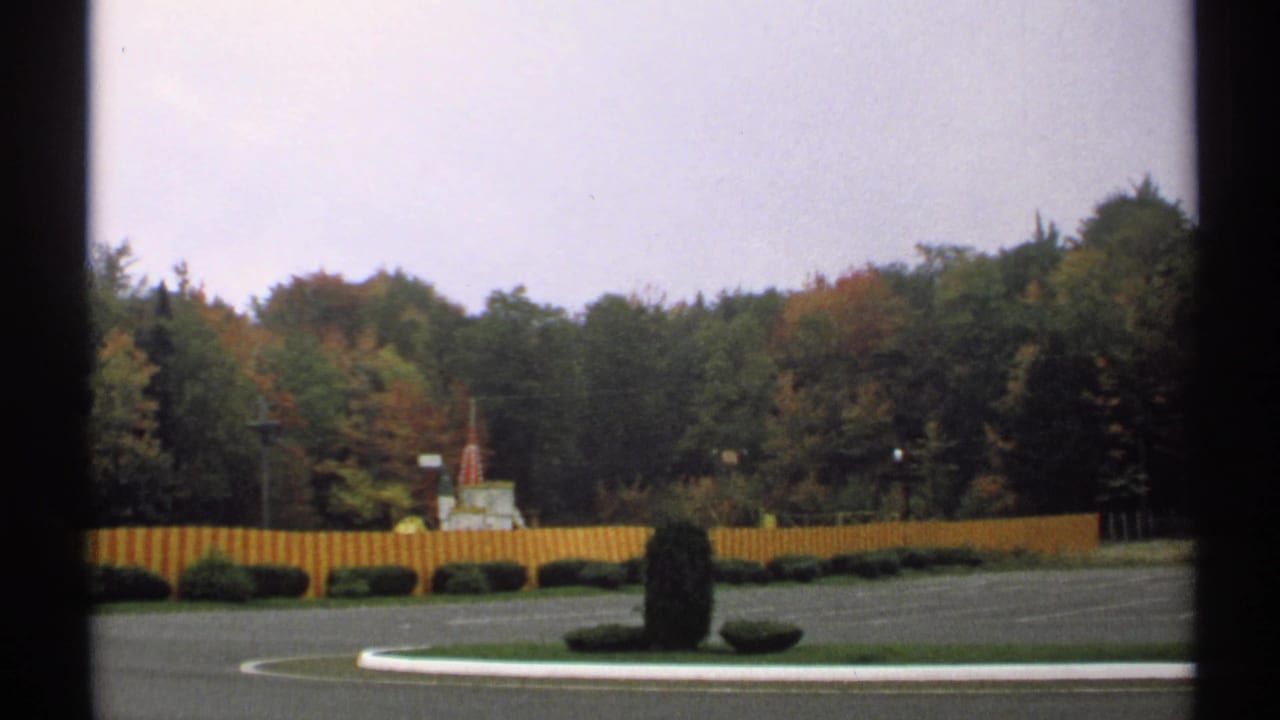 Similar roadside view with fence, shrubs, trees, and cloudy sky.